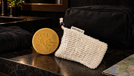 King Rilla round soap and Scrub-a-Dude pouch beside a toiletry bag on a dark hotel vanity.