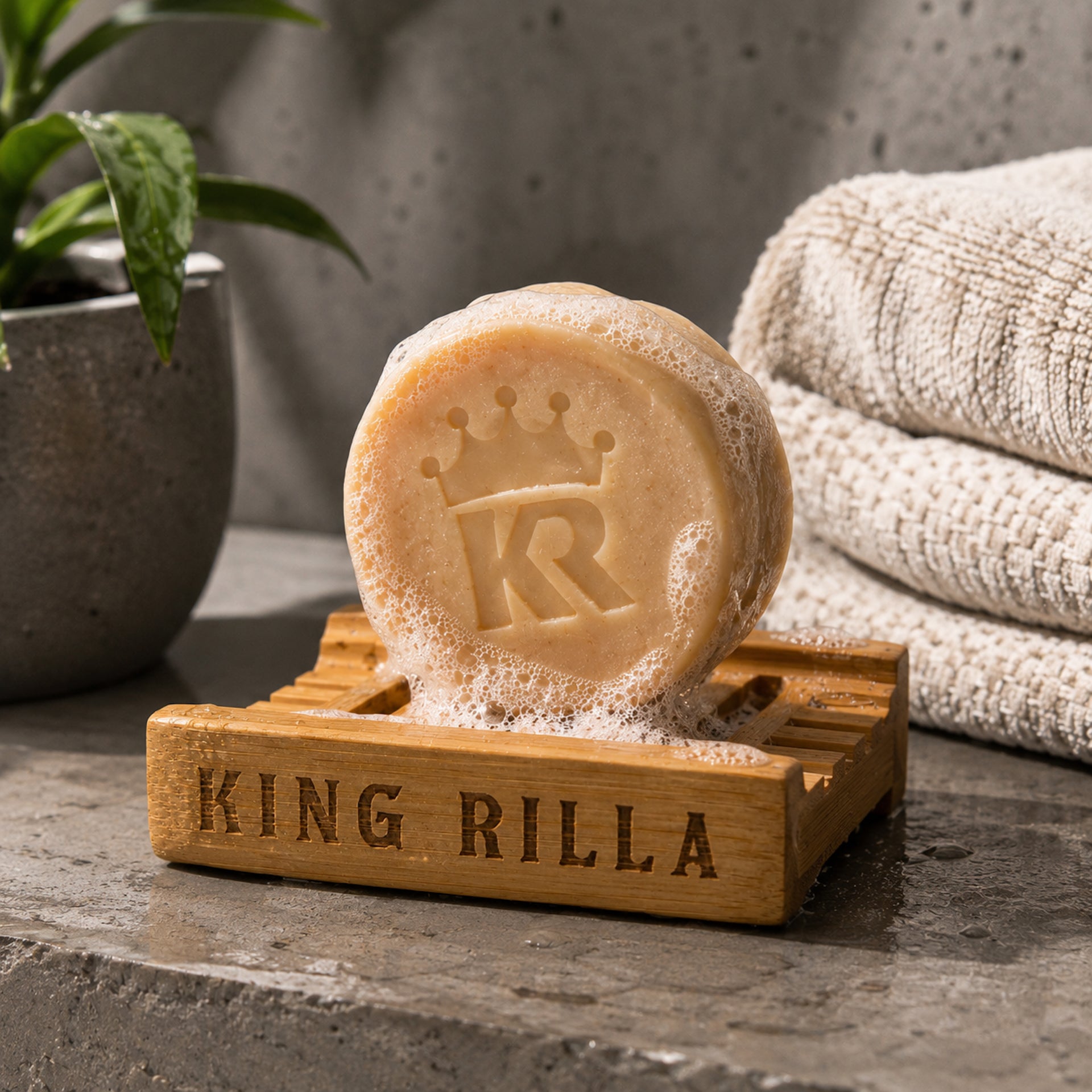 Bar of soap with 'KR' logo on a wooden holder labeled 'King Rilla', with towels and a plant in the background.