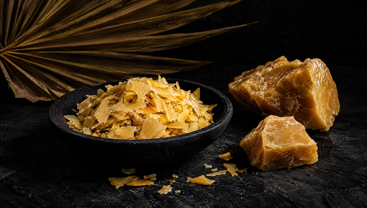 Golden carnauba wax flakes and a fan-palm leaf against charcoal stone.